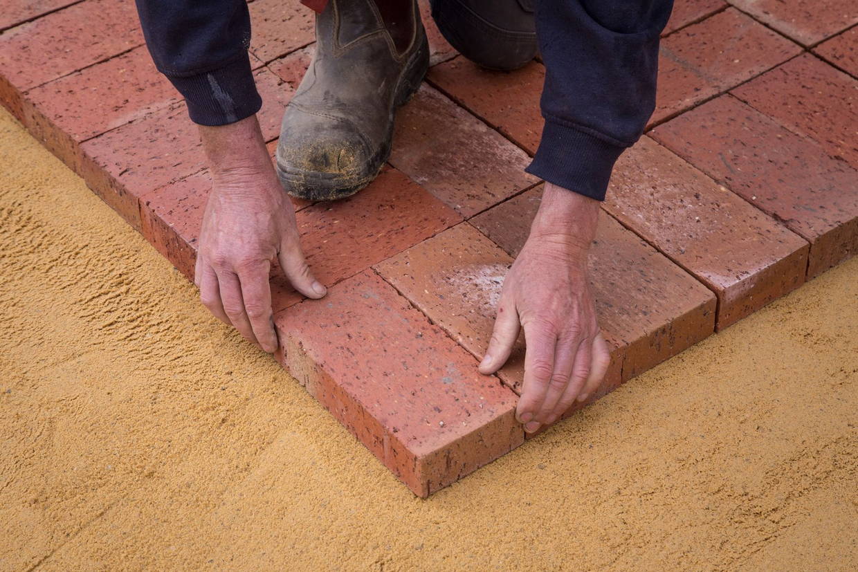Licensed stock photograph: two hands setting a clay paver down onto a screeded bed of sand at the edge of a finished course.